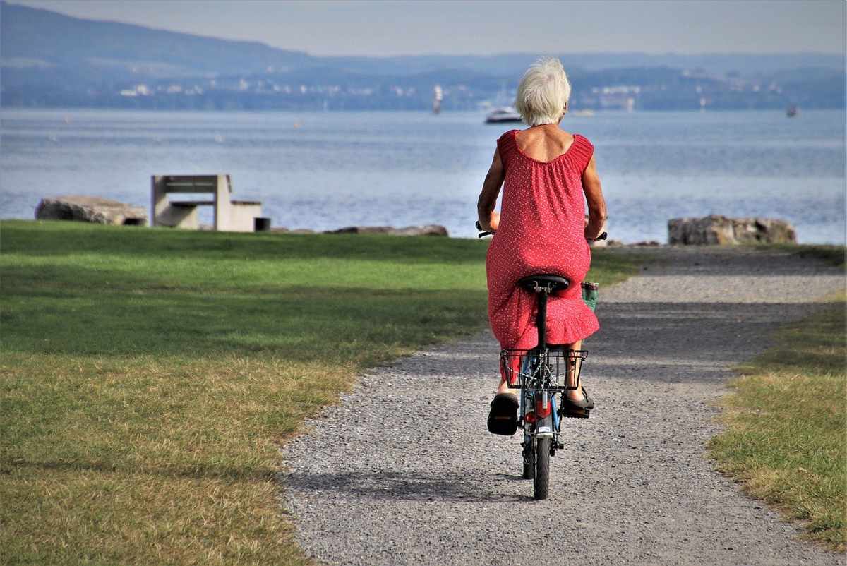 Femme en robe rouge pédale un vélo sur un chemin de gravier près d'un lac avec des montagnes en arrière-plan
