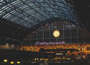 Intérieur d'une gare avec un toit en verre, une horloge, des lumières et des inscriptions